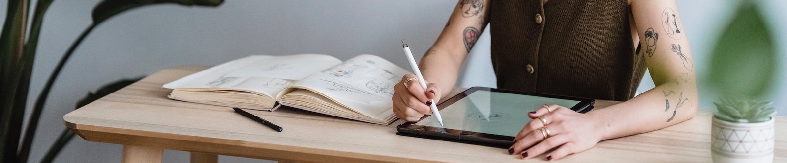 Woman with tattoos on her arms drawing on her Ipad on a desk with a book next to her for inspiration