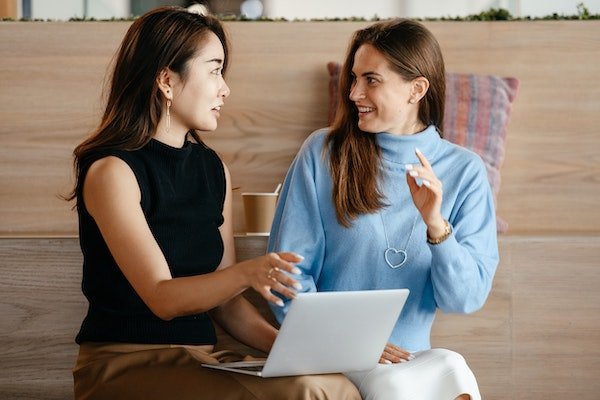 Two women colleagues at work talking. One is dressed in a black sleeveless blazer and tan pants, the other in a baby blue turtleneck and white skirt