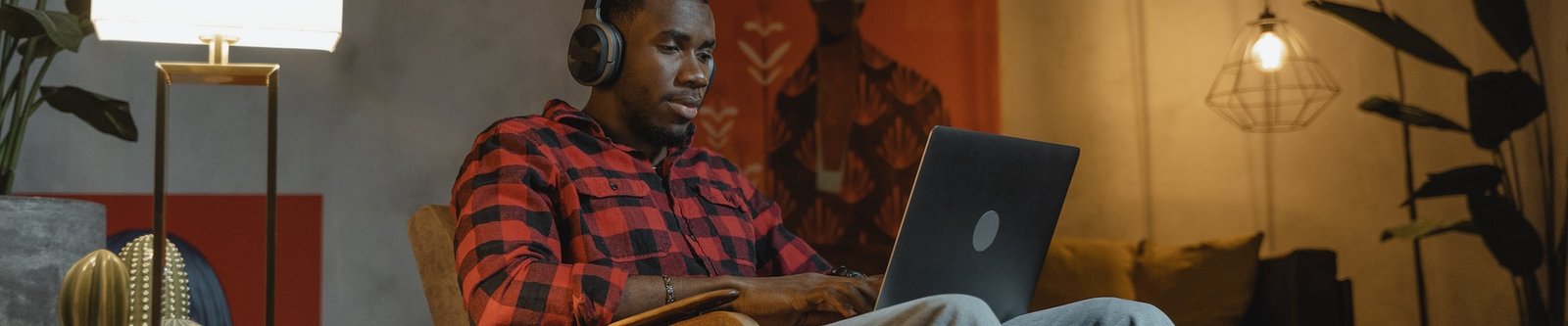 Man in red and black flannel shirt and headphones working in his living room on his laptop